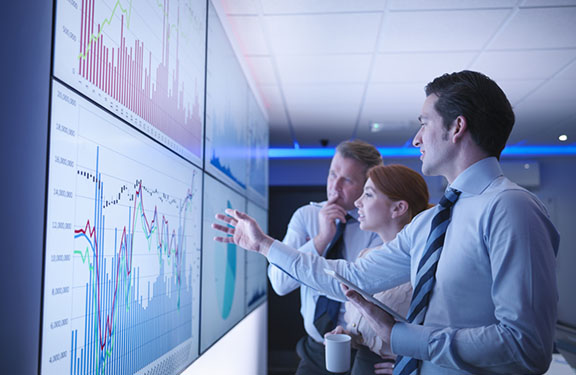 Three professionals in business attire reviewing charts and graphs on a large digital wall display in an office setting.