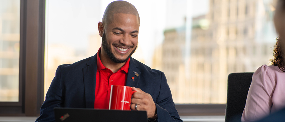 A young man in a suit smiles and holds a coffee mug as he looks at a laptop