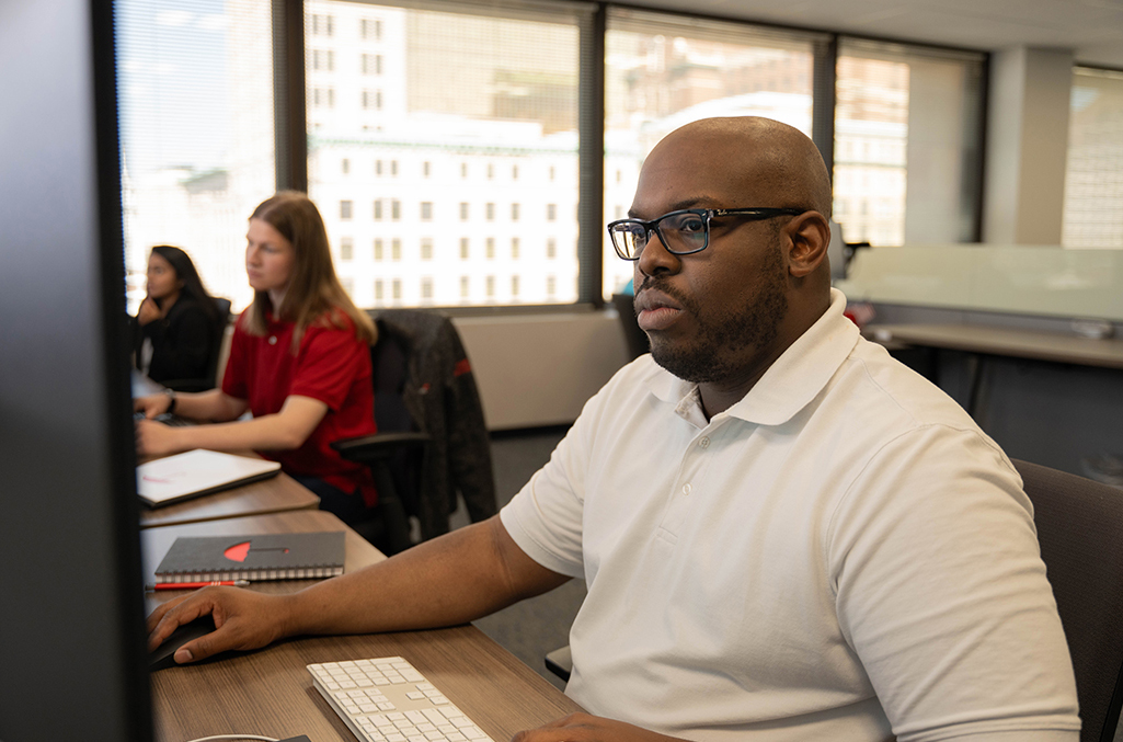 A man with glasses sits at a desk in an office looking at a computer