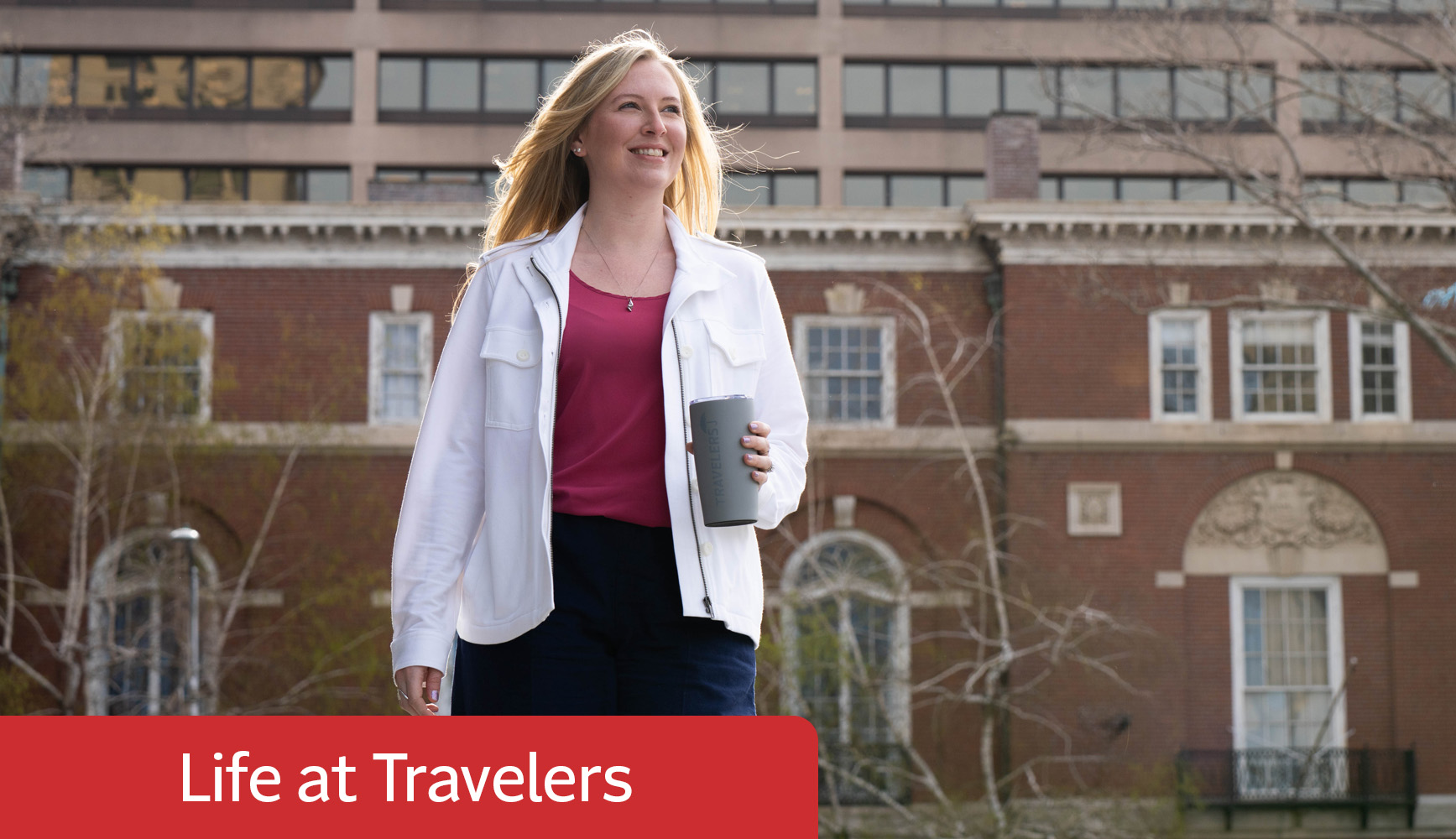 A person walks outdoors holding a tumbler, wearing a white jacket over a red top and dark pants. A large brick building with windows and decorative stonework is in the background, set in an urban environment with trees. A red banner with 'Life at Travelers' is at the bottom of the image