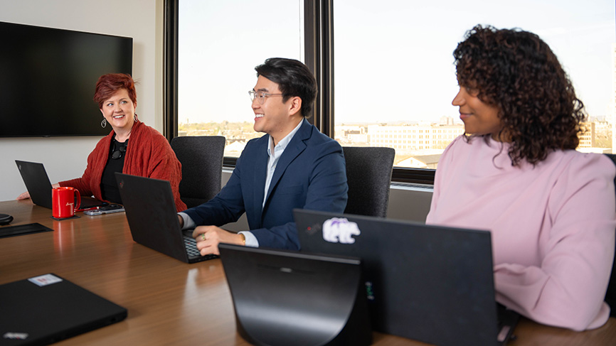 Three colleagues smiling in a meeting room with laptops.