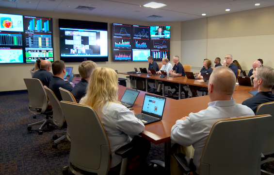 Travelers catastrophe response team in a conference room, reviewing dashboards and video calls on large wall screens during a coordination meeting.