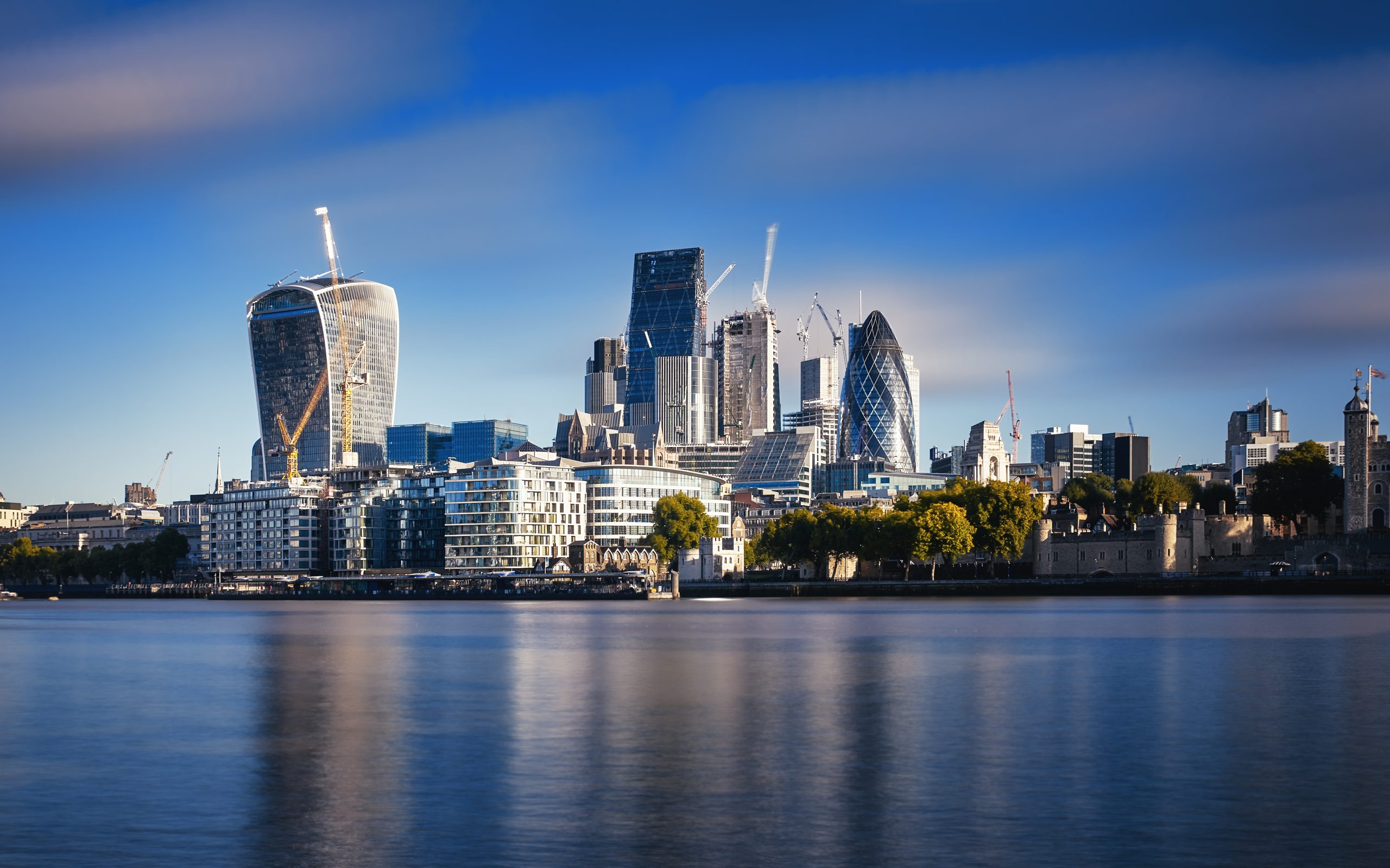 A panoramic view of London's financial district, the City of London, taken from across the River Thames during the golden hour. The skyline features prominent skyscrapers, including the 'Walkie Talkie' building with its unique curved design on the left and the 'Gherkin' with its diamond-patterned glass exterior on the right. The calm river reflects the buildings, and construction cranes indicate ongoing development. The sky is a gradient of blue with scattered clouds, enhancing the dramatic urban landscape.