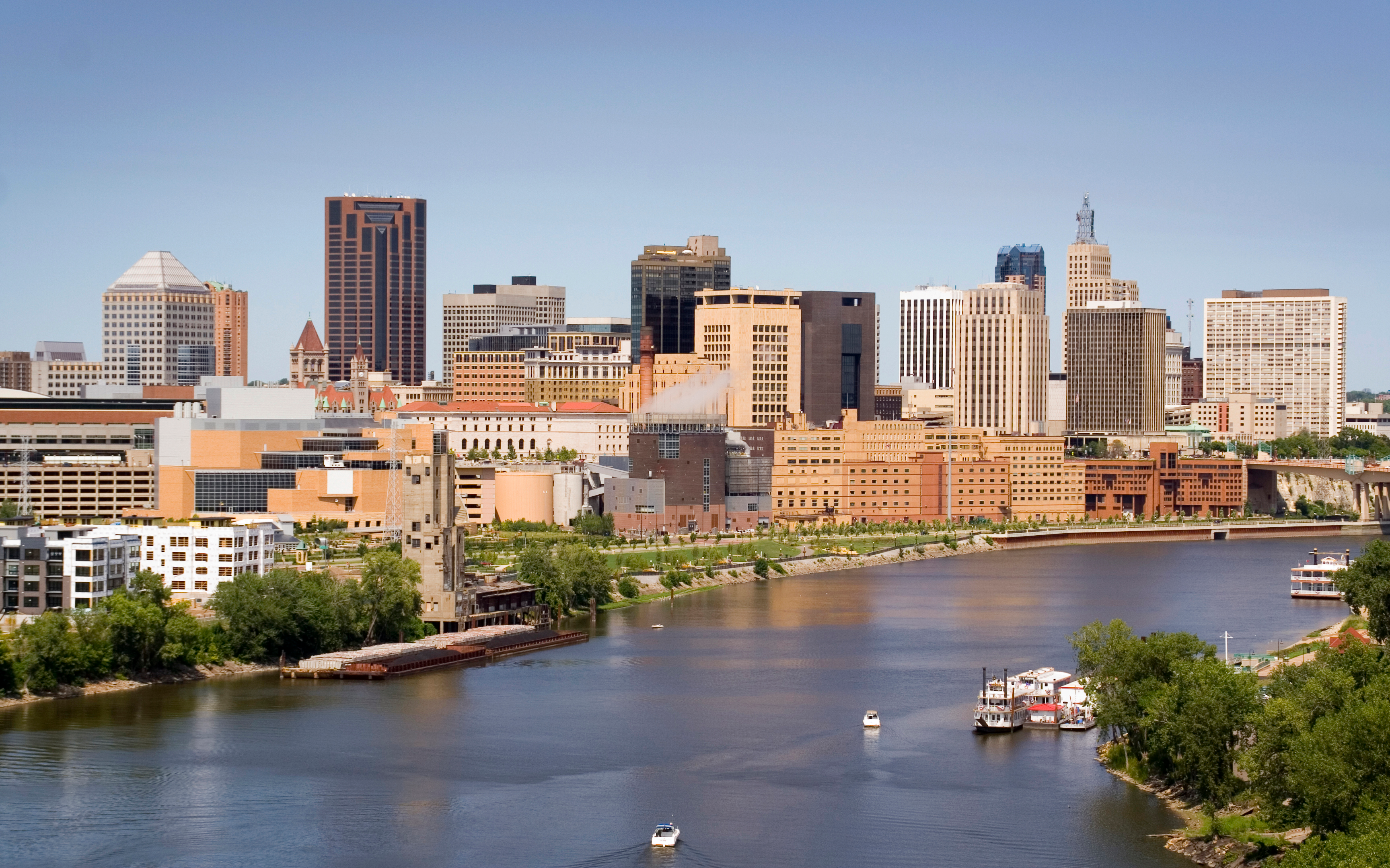 An urban skyline of downtown St. Paul, Minnesota, viewed from across a river. The scene features a variety of modern high-rise buildings and skyscrapers, with a mix of glass and concrete architecture. In the foreground, a wide river is visible, with several boats, including a riverboat or ferry, navigating the water. The riverbanks are lined with trees and some lower-rise buildings. The image is captured during golden hour, casting a warm glow on the buildings against a clear blue sky, highlighting the contrast between the city's development and the natural waterway.