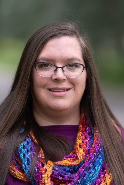 A woman with long, straight brown hair and glasses is smiling warmly. She is wearing a purple top and a vibrant, multicolored crocheted or knitted scarf with bright hues of blue, purple, orange, and yellow. The background is softly blurred, suggesting she is outdoors with greenery or trees behind her. The lighting is natural, giving the photo a professional quality.