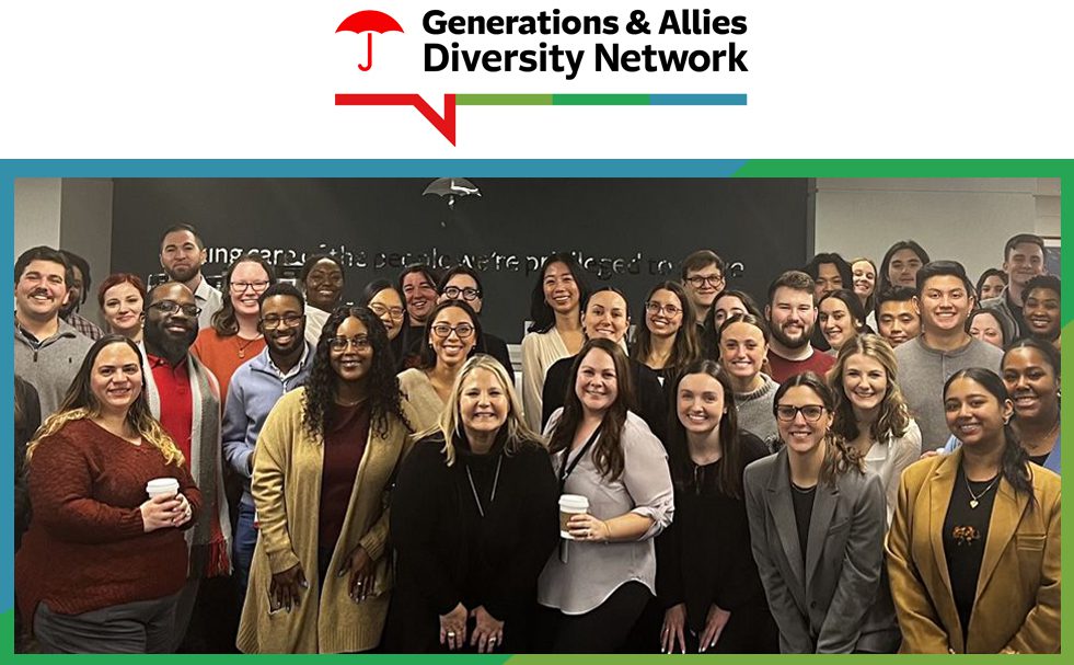 Group photo of diverse professionals at a corporate event, associated with the Generations & Allies Diversity Network. Approximately 30-40 people of various ages, ethnicities, and backgrounds are dressed in business casual attire, smiling and engaged. The logo for the network, featuring a red umbrella icon and colorful horizontal bar, is displayed at the top. The setting is an office environment with a dark wall in the background.
