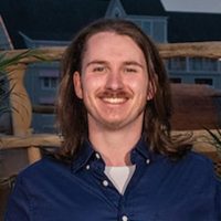 Casual professional portrait of Trentin Louik from Travelers Small Commercial Service & Field Operations. He has shoulder-length brown hair and a mustache, displaying a friendly, approachable smile. He wears a dark blue button-up shirt over a light-colored t-shirt, photographed against a colorful illustrated or animated background