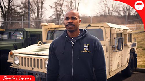 Man in a dark U.S. Marine Corps hoodie standing confidently in front of military vehicles, framed by red branding elements and a “Travelers Careers” graphic in the corner.