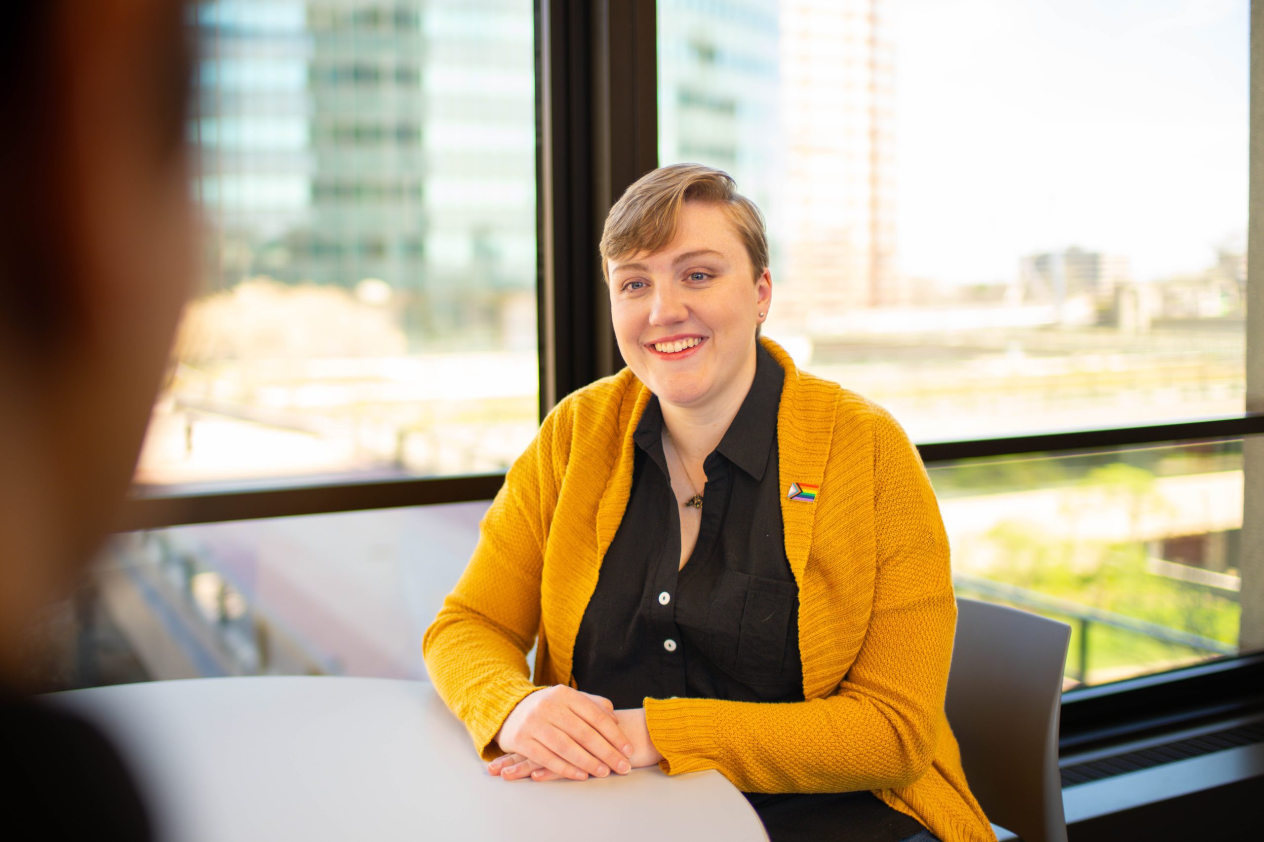 20230411_TRAV_SYMPH_Day2_Shot_5_4288 A professional woman with short blonde hair sits at a white table, smiling at the camera. She's wearing a black button-up shirt under a bright yellow cardigan sweater with a small pin on the lapel. Large windows behind her show a blurred urban cityscape with buildings. The setting appears to be a modern office or conference room with natural lighting.
