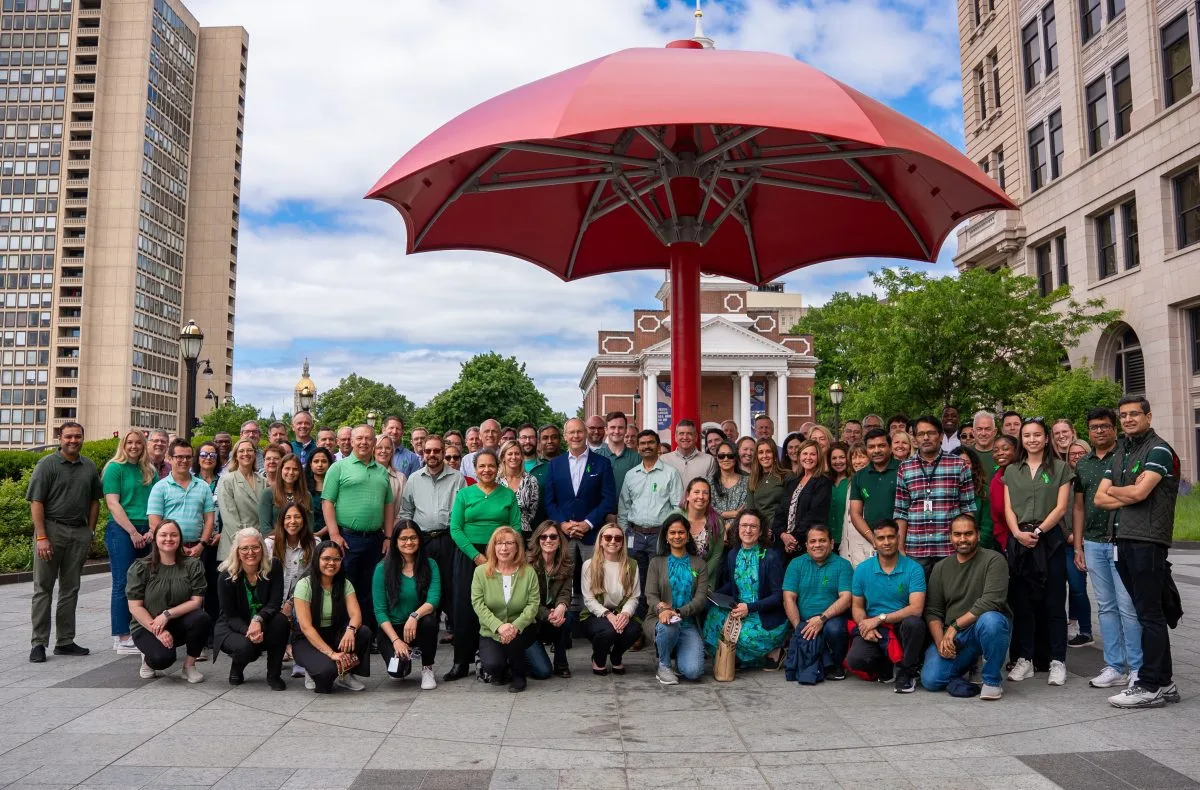 Large group photo of approximately 60-70 people gathered under a red umbrella sculpture in an urban plaza, dressed in coordinated green, blue, and teal business casual attire.