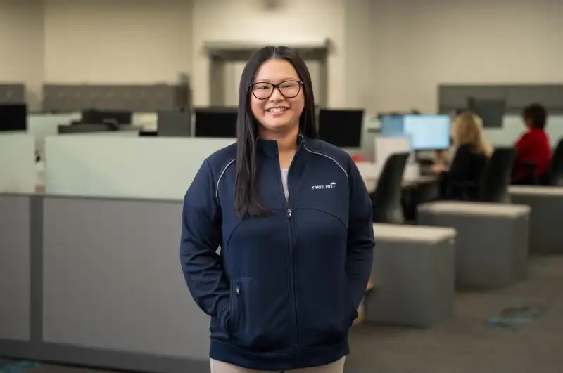A smiling woman with long dark hair and glasses stands in a modern office environment, wearing a navy blue zip-up jacket. Behind her are gray office cubicles with computer workstations, and other employees can be seen working in the background.