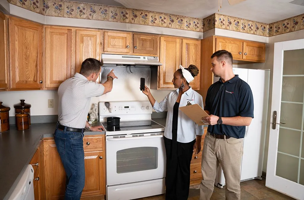 Three people conducting a home inspection in a kitchen with wood cabinets. One inspector on the left examines the range hood while holding a tool, a woman in the center points upward wearing a white shirt and cap, and a male inspector on the right holds a clipboard. The kitchen features honey-colored wood cabinets, white appliances, dark countertops, and decorative wallpaper border.