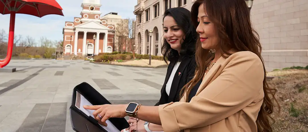 Two women sit outside in a city square and look at a notepad together