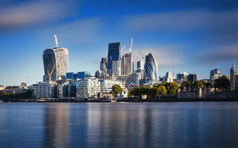 A panoramic view of London's financial district, the City of London, taken from across the River Thames during the golden hour. The skyline features prominent skyscrapers, including the 'Walkie Talkie' building with its unique curved design on the left and the 'Gherkin' with its diamond-patterned glass exterior on the right. The calm river reflects the buildings, and construction cranes indicate ongoing development. The sky is a gradient of blue with scattered clouds, enhancing the dramatic urban landscape.
