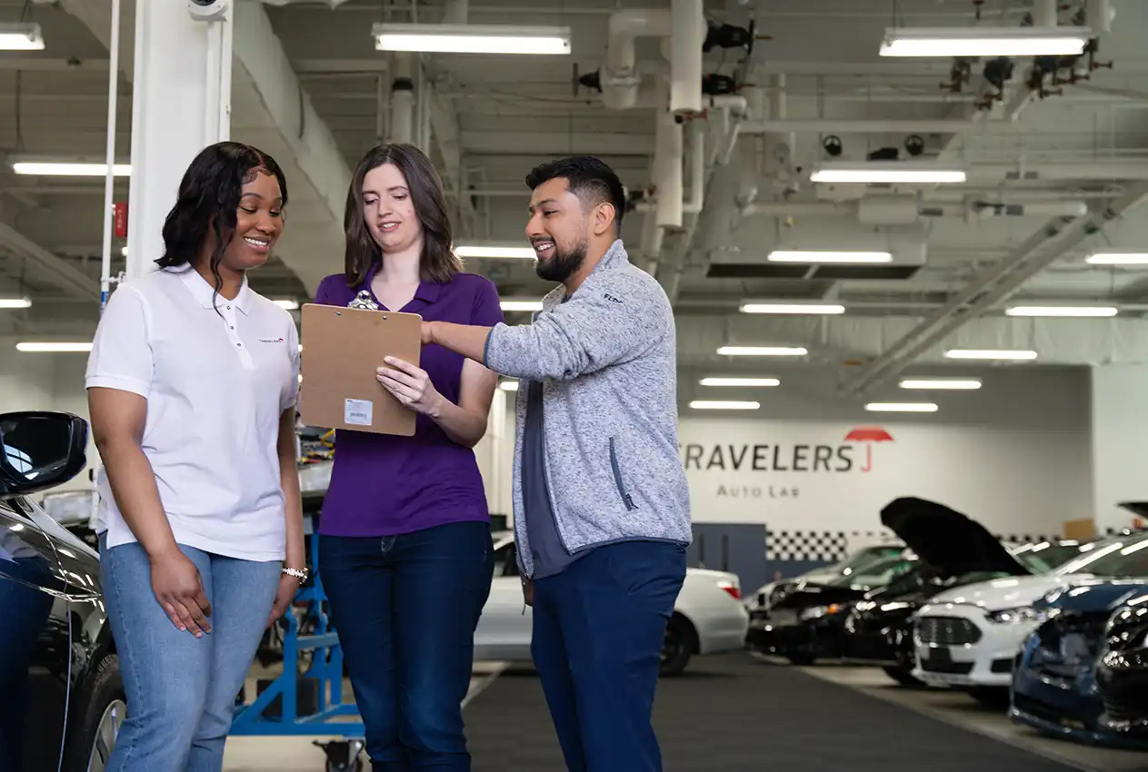 Three people stand together in an automotive service facility with an industrial ceiling and fluorescent lighting. They are reviewing a document or clipboard. On the left is a woman in a white polo shirt and jeans, in the middle is a woman in a purple top and jeans, and on the right is a man in a gray sweater and jeans. A 'TRAVELERS' sign is visible on the wall, and several cars are in the background.