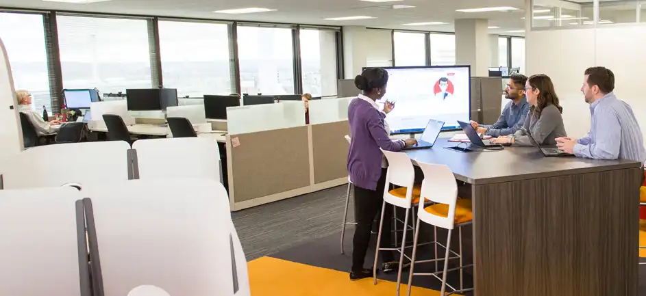 sg-intern-intro-optimized Three people sit and one stands at a tall table in an office, looking at a screen together